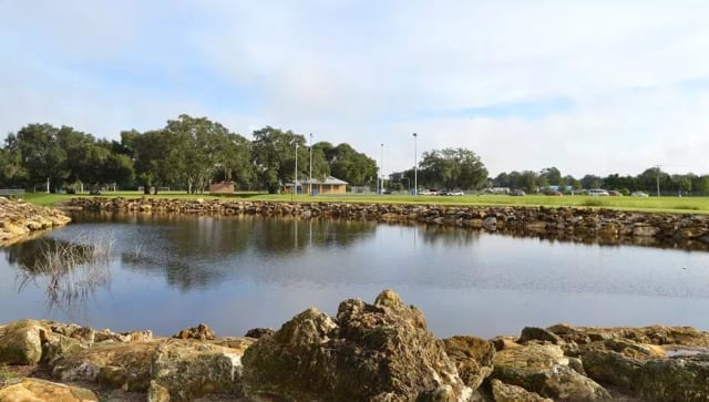 Rocky shoreline at LaBelle Civic Park with sports fields and trees in the background.